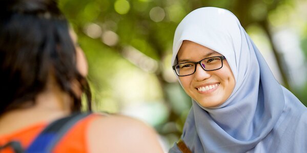 A smiling girl in a light blue hijab greets her friend.