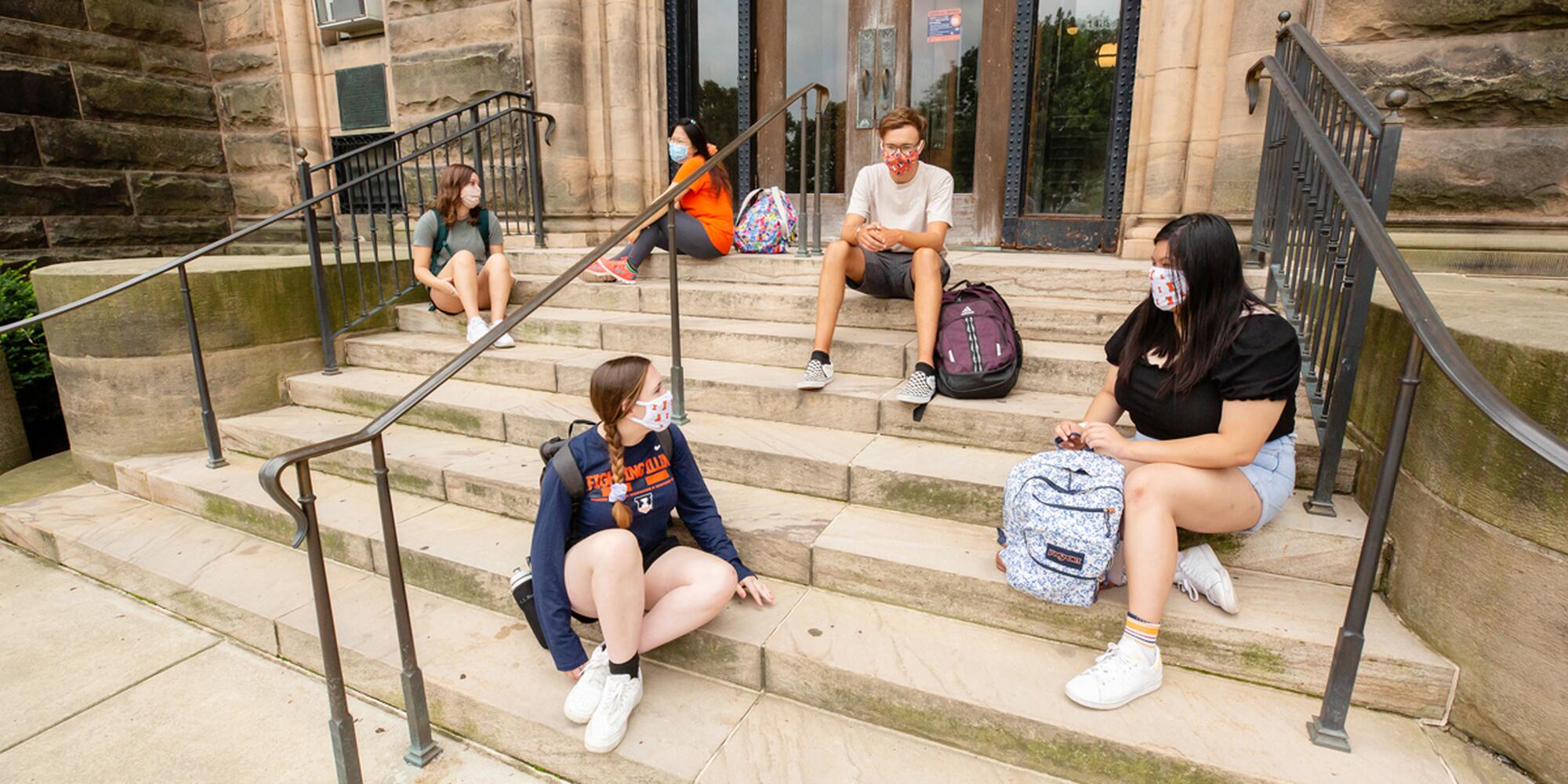 Masked students sit apart from each other on a set of steps.