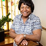 A photo of Mary Ellerbe. She is pictured from the waist up in a patterned black and white dress, and is smiling brightly at the camera.