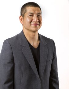 An image of Eric Tang. He is visible from the waist up, and wears a gray blazer over a black shirt. His hair is cut short. He smiles at the camera.
