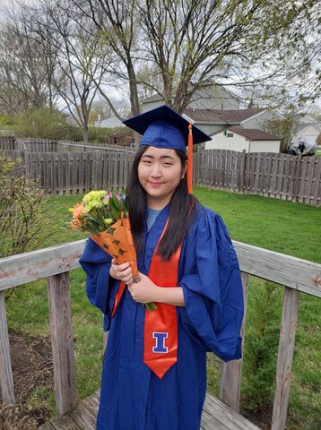 Photo of Julia Son in her graduation regalia. She is smiling at the camera and holding a bouquet of yellow flowers.