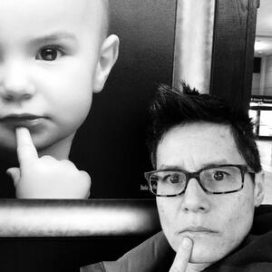A black and white photo of Dr. Fiona Ngo. She is standing next to a large poster of a baby holding a finger to its chin, and is posed next to it in imitation.