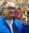 Photo of Dr. Martin Manalansan. He is wearing a blue jacket and is smiling while holding up a bottle of Sriracha Rooster Sauce.