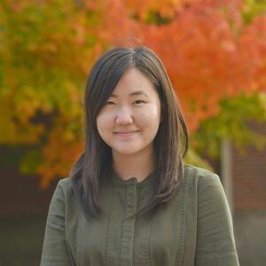 A photo of Jacqueline Yi. She is smiling at the camera and wearing a green shirt. Behind her are autumn trees.
